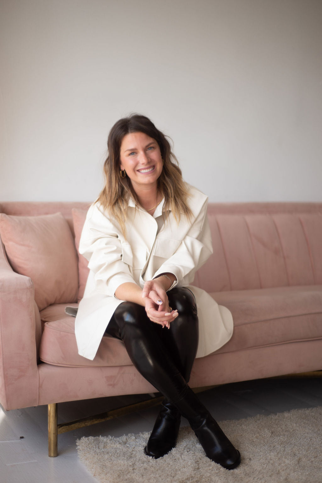Woman sitting on a pink couch wearing a white coat and black leggings.